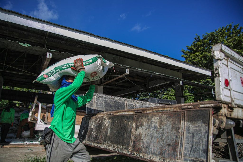 A person carries a bag of fertilizer to a truck outside a warehouse, illustrating hard outdoor labor.